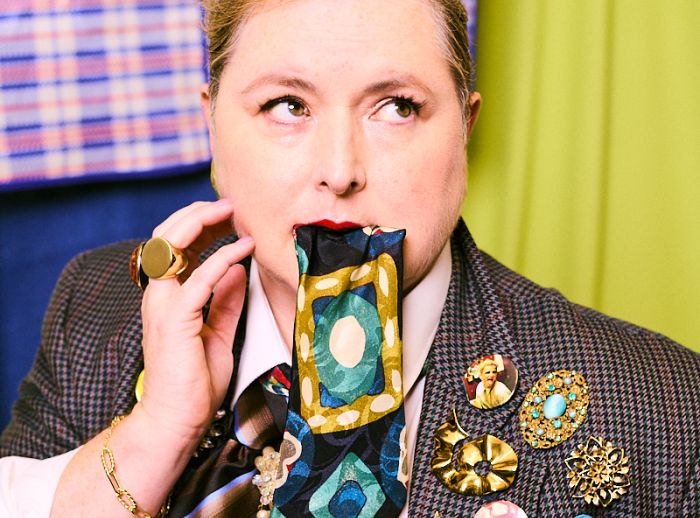 Siobhan McSweeney posing with a tie in her mouth and wearing a vest with many cool badges and broches