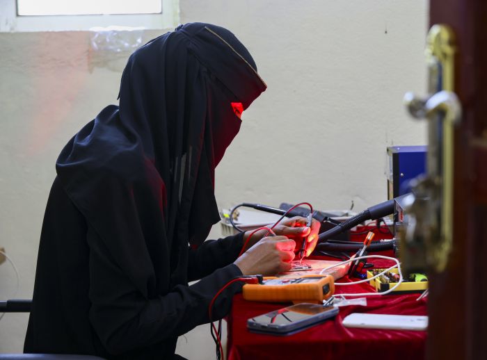 Nahla Abdo repairs an electronic device at a table with tools and components in a naturally lit indoor setting.