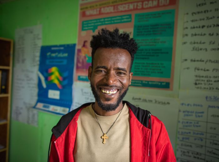 Aberaw Mamo, a nurse, smiles at the camera in a clinic. He wears a red jacket, beige shirt, and a cross necklace. He and his colleagues provide confidential family planning and counselling to young people.