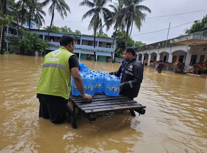 Oxfam workers in floodwater deliver bottled water using a cart in a submerged community.