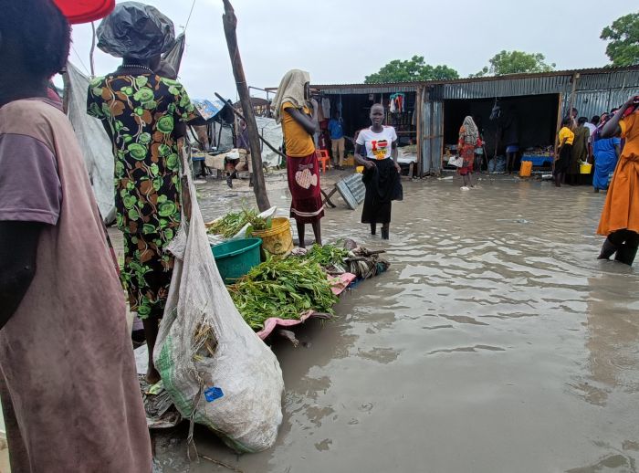 Flooded market in South Sudan with people wading through ankle-deep water and makeshift stalls in the background.