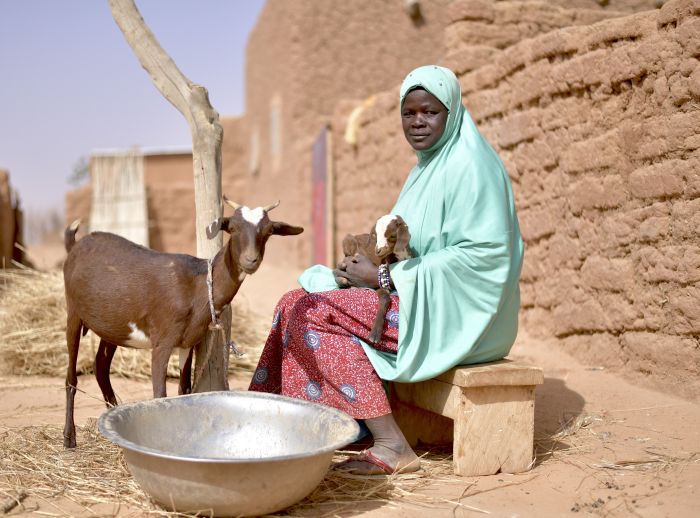 A woman in Ouallam, Niger, sits on a bench holding two goats, with another goat nearby, highlighting the impact of climate change and hunger on her family.