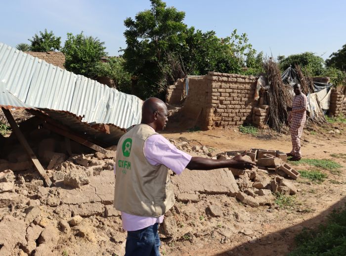 Collapsed brick structure with corrugated metal roof; two people, including one in an Oxfam vest, assess the debris.