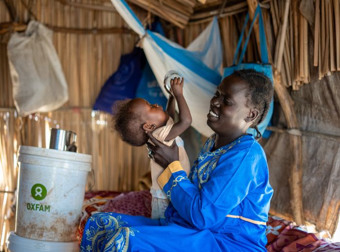 Ayul Ajak, a visually impaired Sudanese refugee mother, playing with her baby inside a makeshift shelter with an Oxfam bucket nearby.