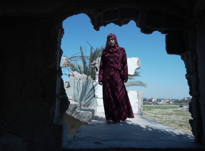 A woman in a red dress stands in a bombed-out building, symbolizing maternal hardship in Gaza, supported by the Power to Choose Programme.