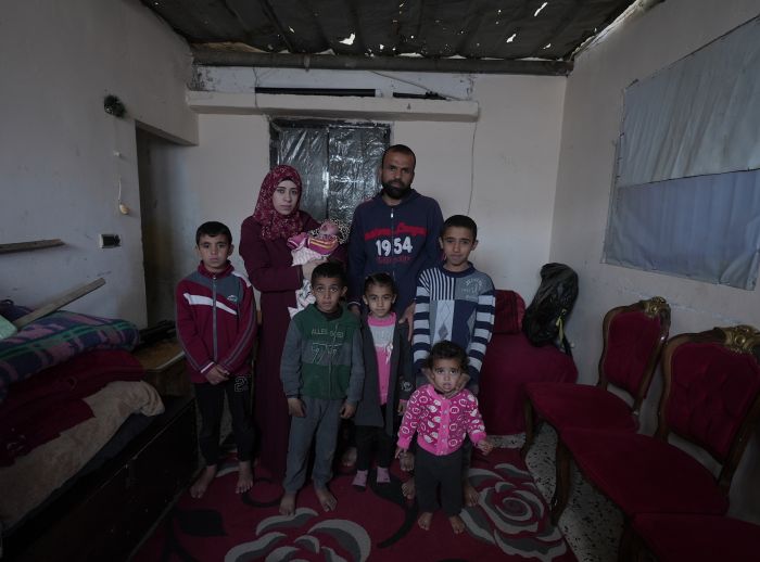 Suhad, her husband, and their children stand in a room with red chairs, a bed, and a damaged ceiling."