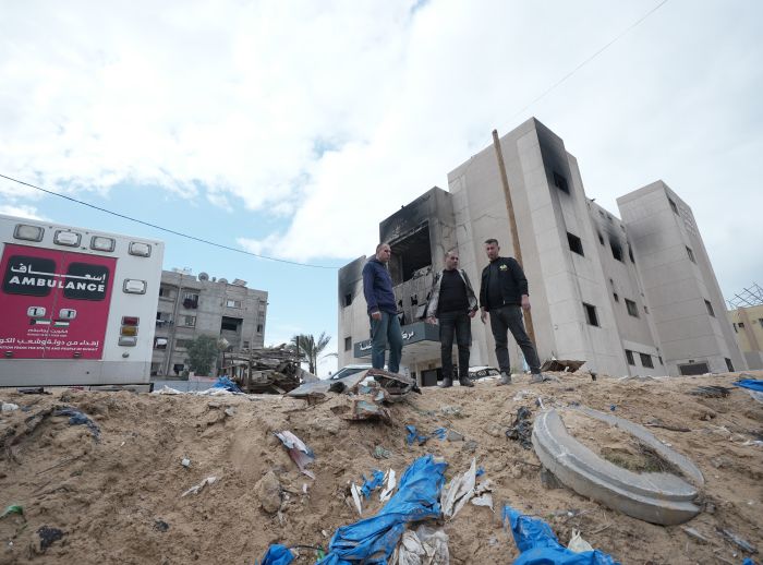 Three men stand surrounded by rubble and an ambulance.