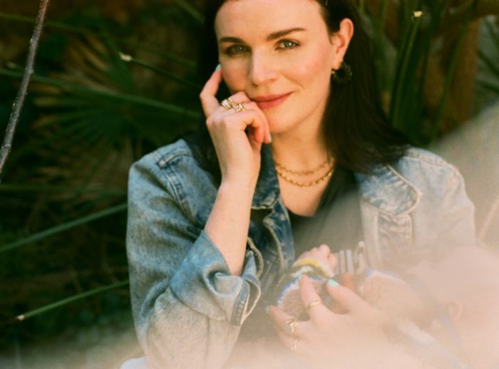 Aisling Bea looks into the camera holding her baby, wearing a denim jacket.