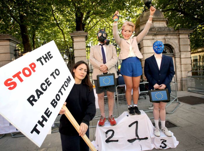 Stunt at Leinster house. Banner reads "Stop the Race to the bottom," 3 people are standing on a podium representing the EU with masks