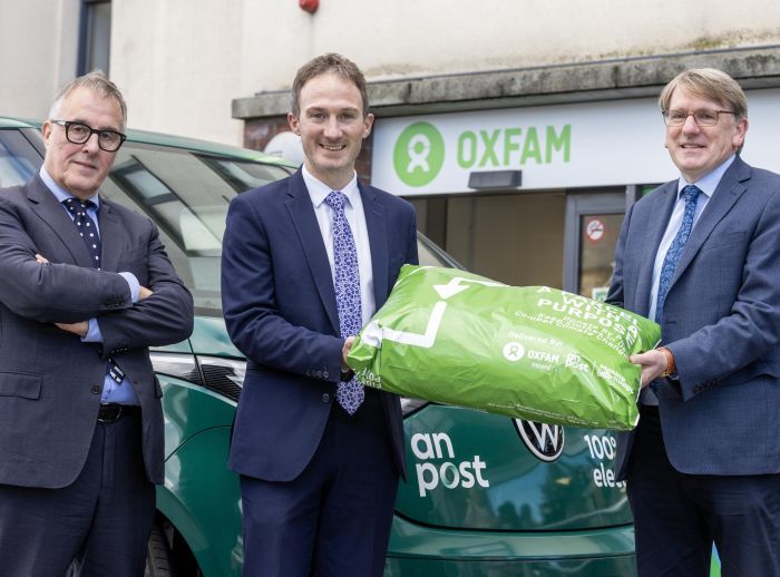 Three men holding a green PostBack package in front of a dark green An Post van and Oxfam store