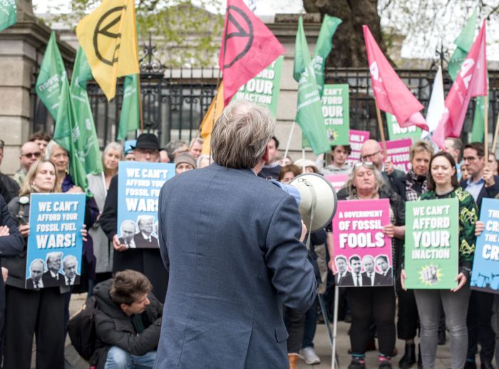 Man wearing suit holds megaphone and speaks to people holding climate placards and flags in front of Dáil Éireann.