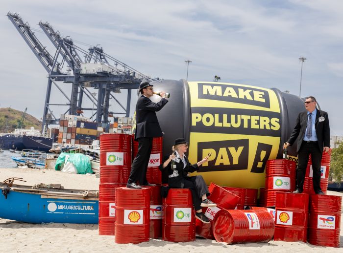Three people in formal suits stand and sit on stacked red oil drums beside a large black inflatable with the words “MAKE POLLUTERS PAY,” set in an industrial port with cranes, cargo containers, and boats in the background.