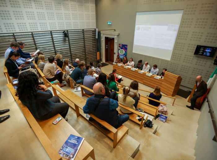 Room with wooden benches with people watching presentation.