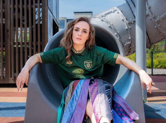 Comedian Emma Doran is sitting on a grey playground slide wearing a tie skirt.