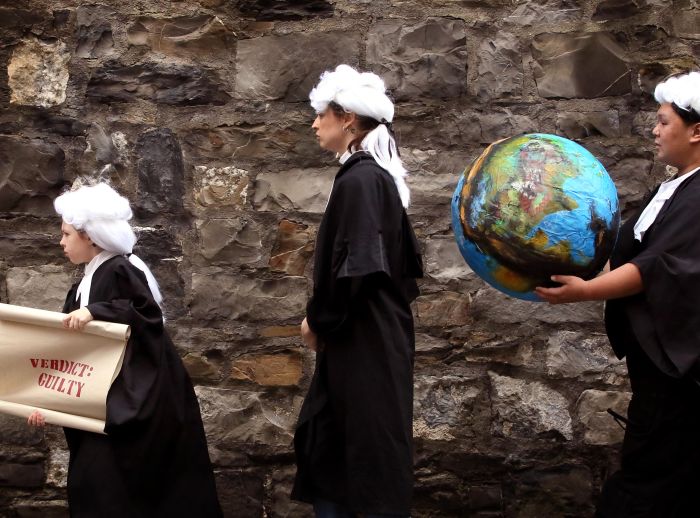Young climate judges holding a scroll saying 'verdict: guilty' and giant globe while wearing robes and wigs.