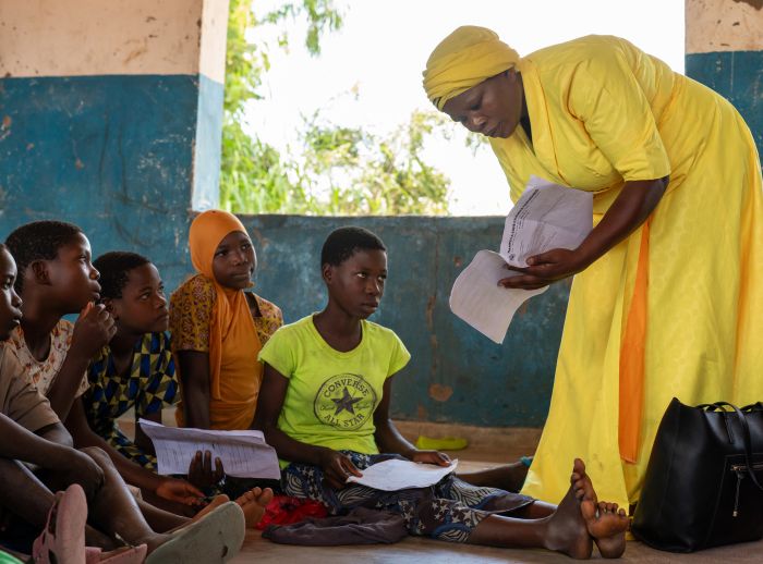 A lady dressed in yellow leans over to show children something on sheets of paper she is holding