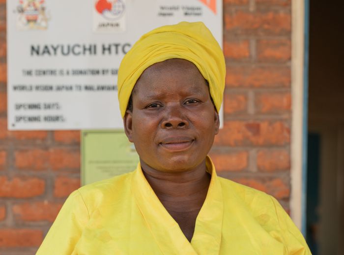 A lady dressed in yellow looks into the camera. Her hands are folded
