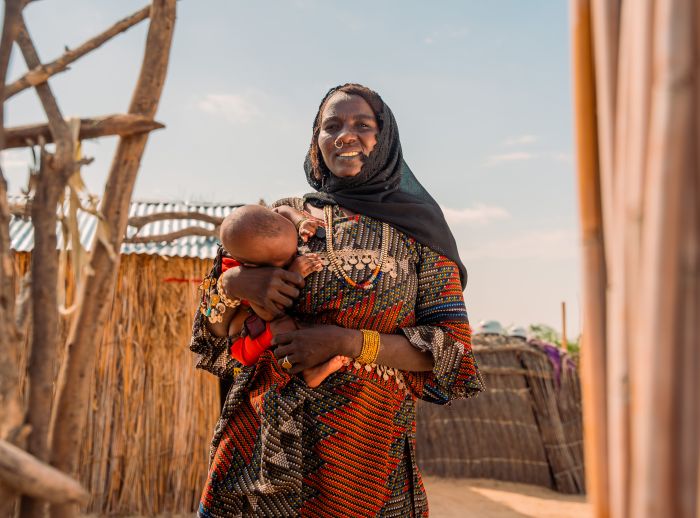 A lady stands holding a baby. She is wearing a dark headscarf, fence structures are around her