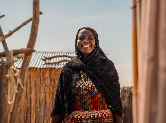 A lady smiles at the camera. She is wearing a black headscarf and colourful dress. There are stick fences around her