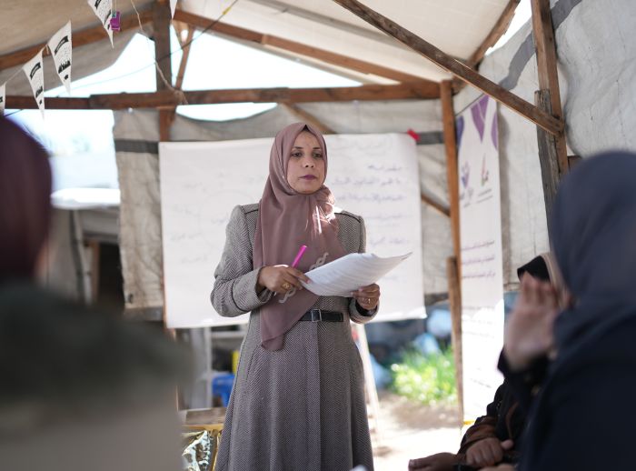 A lady in a pink hijab stands holding sheets of paper in a room with other people listening to her