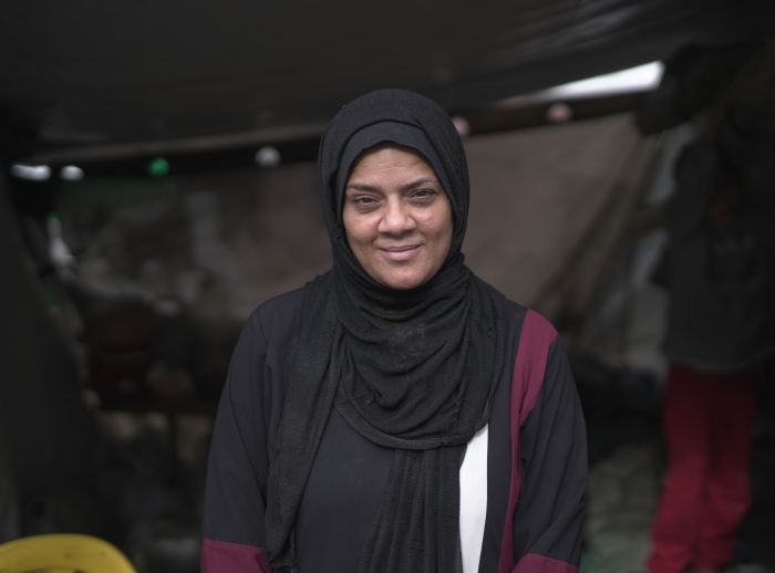A lady in a black hijab smiles at the camera. She is in a makeshift tent camp in Gaza