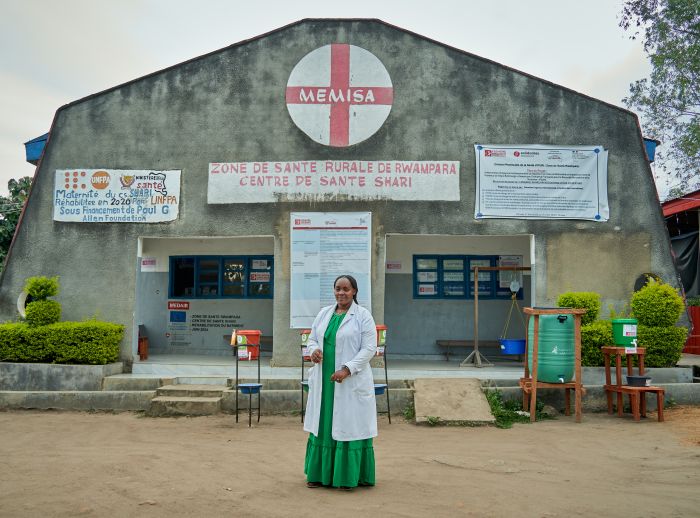 A lady in a green dress and long white coat stands in front of a hospital