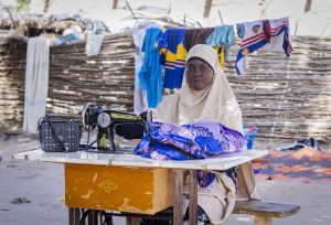 Zahara Seybou Harouna sitting outside, posing while working on sewing clothes with her sewing machine.