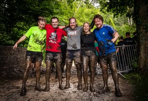 A group of 4 men and a women at the end of the Hell and Back Challenge. They are holding each other side by side, smiling and covered in mud.