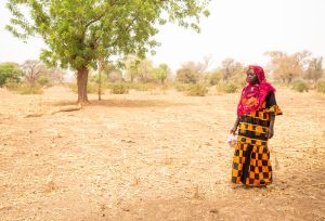 Sanfo Ramata, a 49-year-old farmer in Burkina Faso, stands in a dry field affected by climate change, wearing a colorful dress and red headscarf.