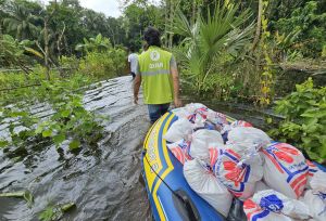 Oxfam staff pull a supply-laden inflatable boat through a flooded area surrounded by greenery.