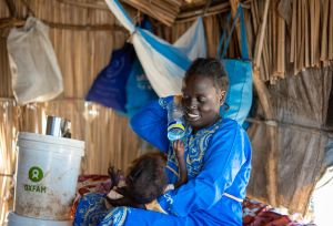 Ayul Ajak, a visually impaired Sudanese refugee mother, breastfeeds her baby inside a makeshift shelter with an Oxfam bucket nearby.