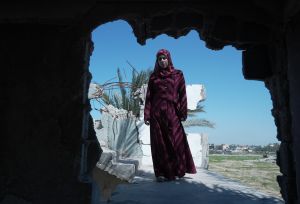 A woman in a red dress stands in a bombed-out building, symbolizing maternal hardship in Gaza, supported by the Power to Choose Programme.