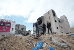 Three men stand surrounded by rubble and an ambulance.