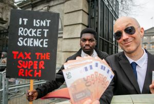 Man dressed up as billionaire holding cash stands infront of protester holding Tax the Super Rich placard