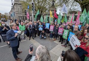 Crowd gathered outside Dáil Éireann with placards while man holds megaphone wearing a suit.