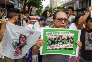A man holding a sign reading “Stop Fossil Fuels – climate chaos” while standing in a crowded street protest with others carrying banners and flags.