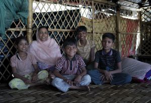 Romida sits with four children inside a bamboo shelter in Cox’s Bazar camp, where the family has lived for seven years after fleeing violence and faces safety concerns in poorly lit areas.