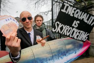 A man with a rocket, money and a cigar and a protester behind him with a sign saying inequality is skyrocketing