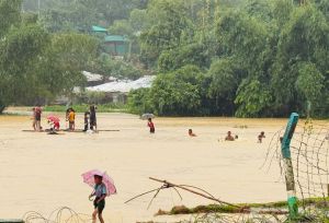 People move through floodwater in Cox’s Bazar after heavy rain, with umbrellas visible near flooded shelters and trees.