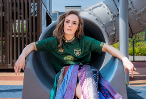 Comedian Emma Doran is sitting on a grey playground slide wearing a tie skirt.