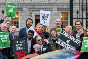 A group of people stand outside Leinster house holding signs that say Wealth Tax Now