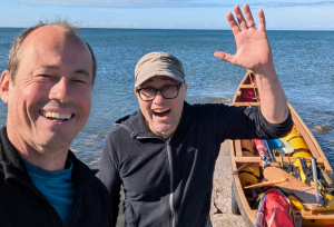 Johan Hoving smiling on the left and Rich Ensoll waves on the right beside their canoe.