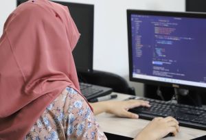 A lady in a pink headscarf works on a computer. Her hands are placed on the keyboard, the screen of the computer is blue