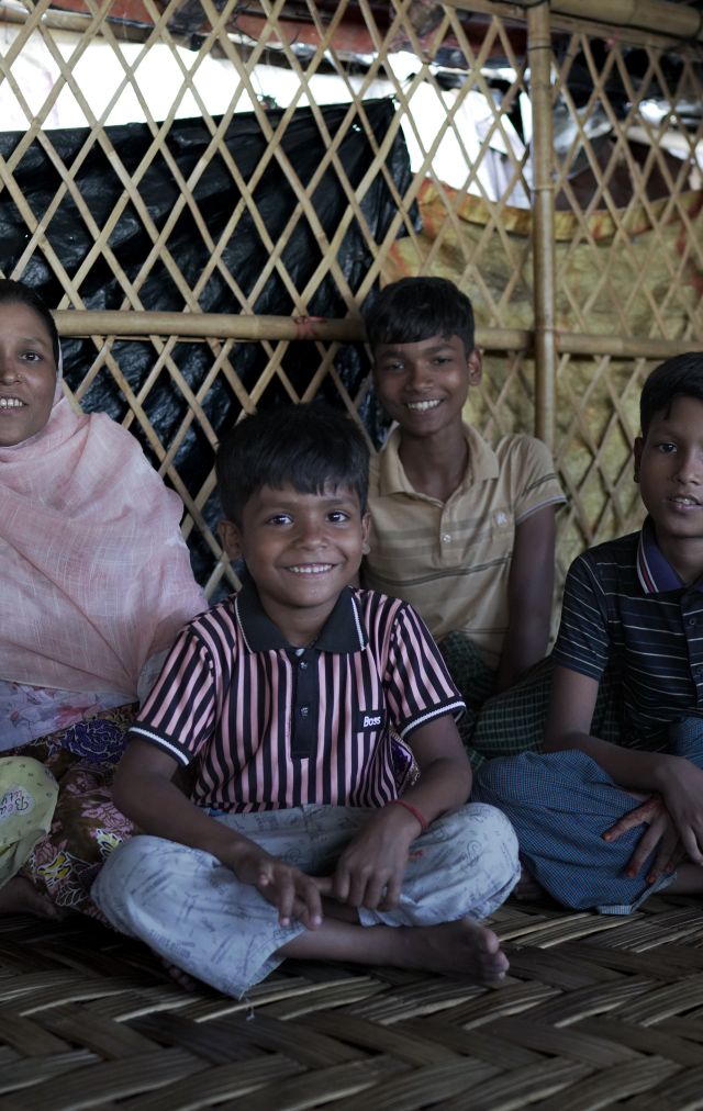 Romida sits with four children inside a bamboo shelter in Cox’s Bazar camp, where the family has lived for seven years after fleeing violence and faces safety concerns in poorly lit areas.