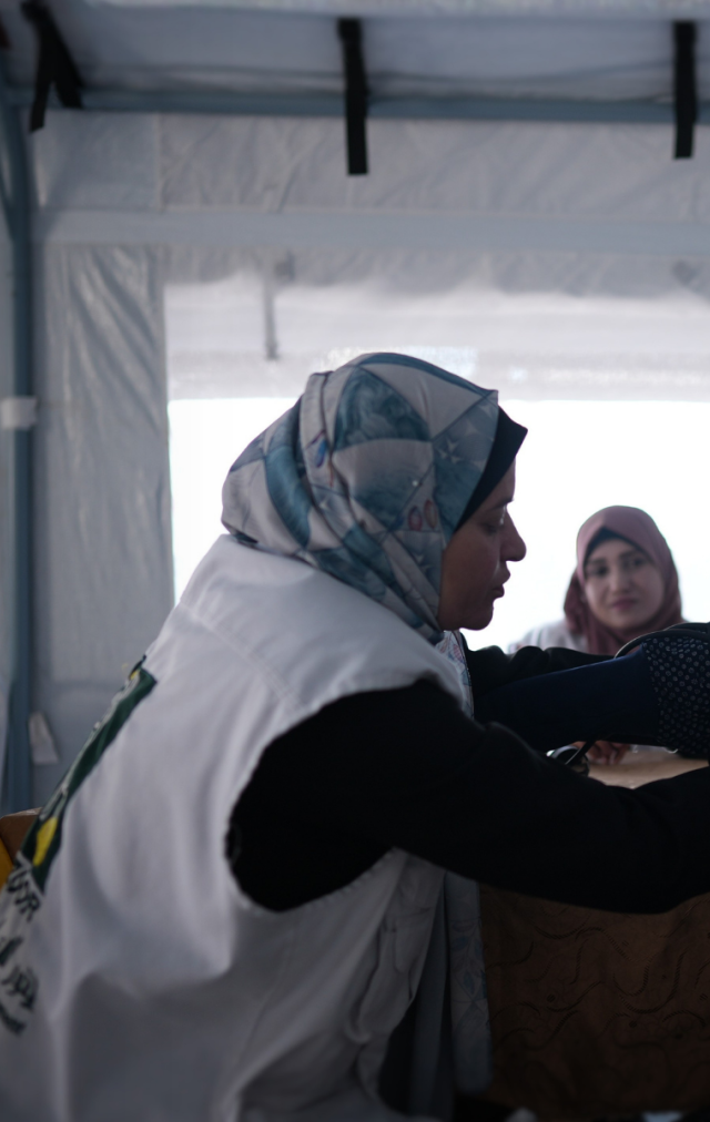 Three women sit in a white room. One is a doctor, she is taking the blood pressure of another woman in the foreground