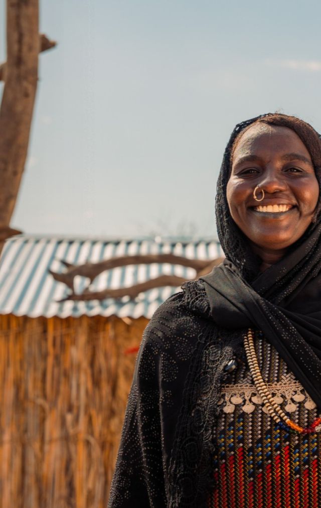 A lady in a black hijab smiles at the camera. There are stick structures and a fence in the background