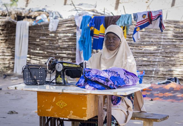 Zahara Seybou Harouna sitting outside, posing while working on sewing clothes with her sewing machine.