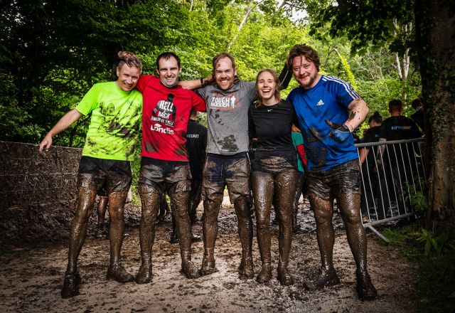 A group of 4 men and a women at the end of the Hell and Back Challenge. They are holding each other side by side, smiling and covered in mud.