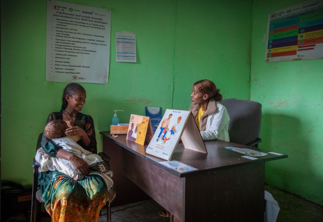 A young mother with her baby speaks to a smiling nurse in a green-walled clinic in Ethiopia, with health posters and educational materials visible.