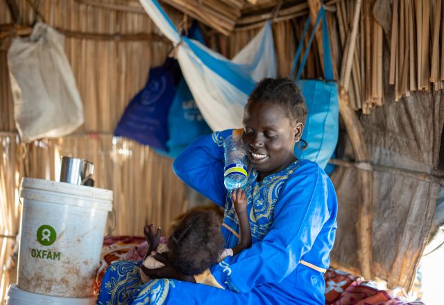 Ayul Ajak, a visually impaired Sudanese refugee mother, breastfeeds her baby inside a makeshift shelter with an Oxfam bucket nearby.